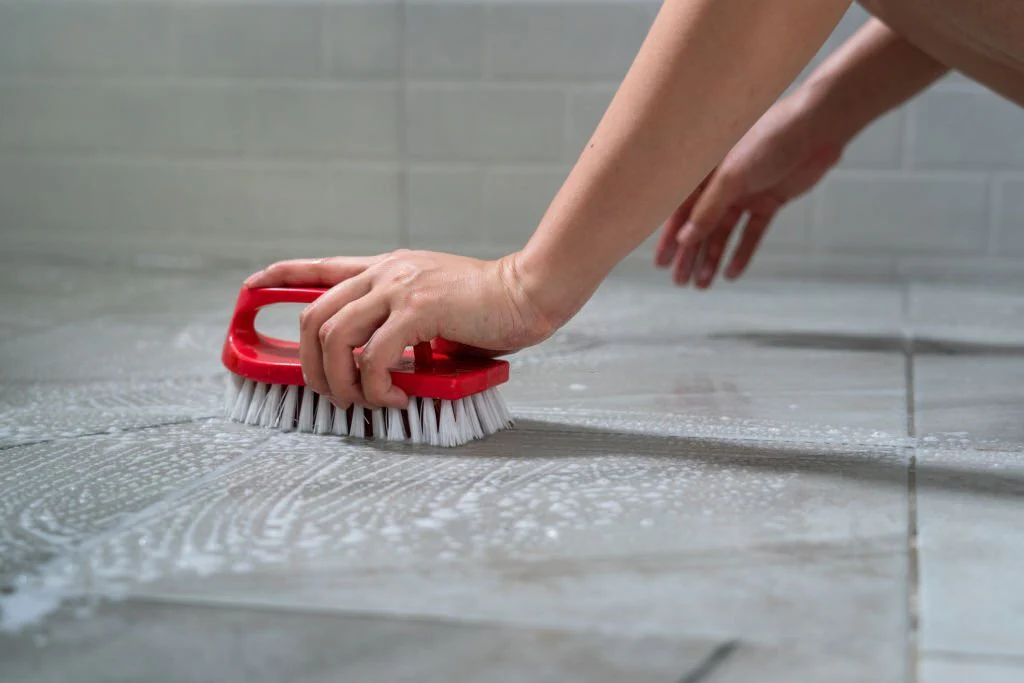 A soft nylon brush gently scrubbing a textured porcelain tile floor near a kitchen cabinet with a small amount of cleaner, realistic home photo