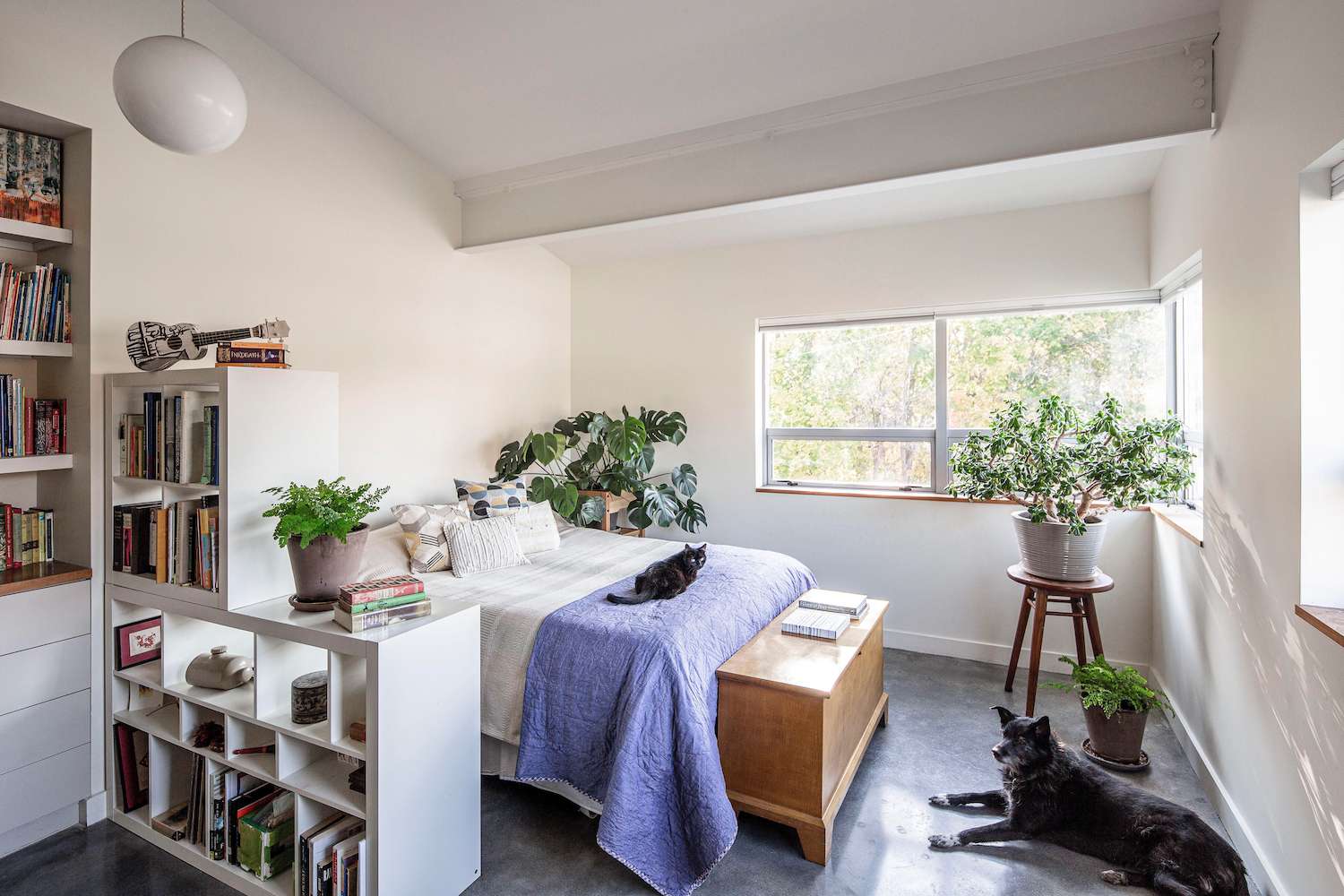 A tall bookcase placed against a bedroom wall behind a bedside table, filled with books and woven baskets, styled in a cozy renter apartment, real photo