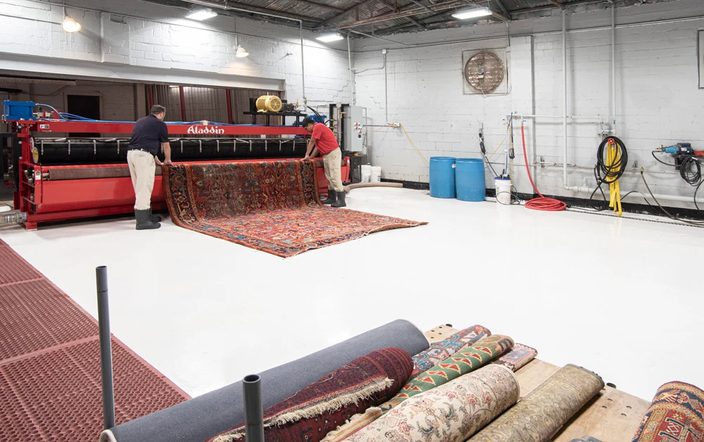 A technician at a rug washing facility rinsing a wool area rug on a wash floor with controlled drainage and drying equipment in the background