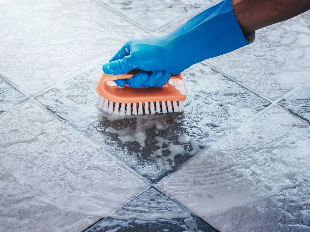 A textured ceramic tile bathroom floor being gently scrubbed with a small soft-bristle brush along the grout lines, close-up photo in warm indoor light