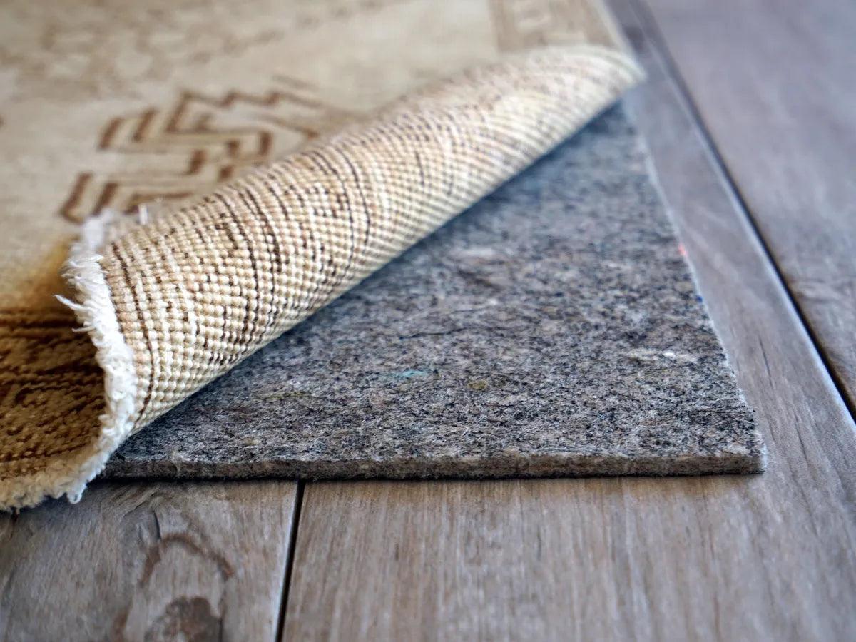 A thick wool area rug partially folded back to show a dense felt rug pad underneath on a bedroom floor, real-life home interior photo