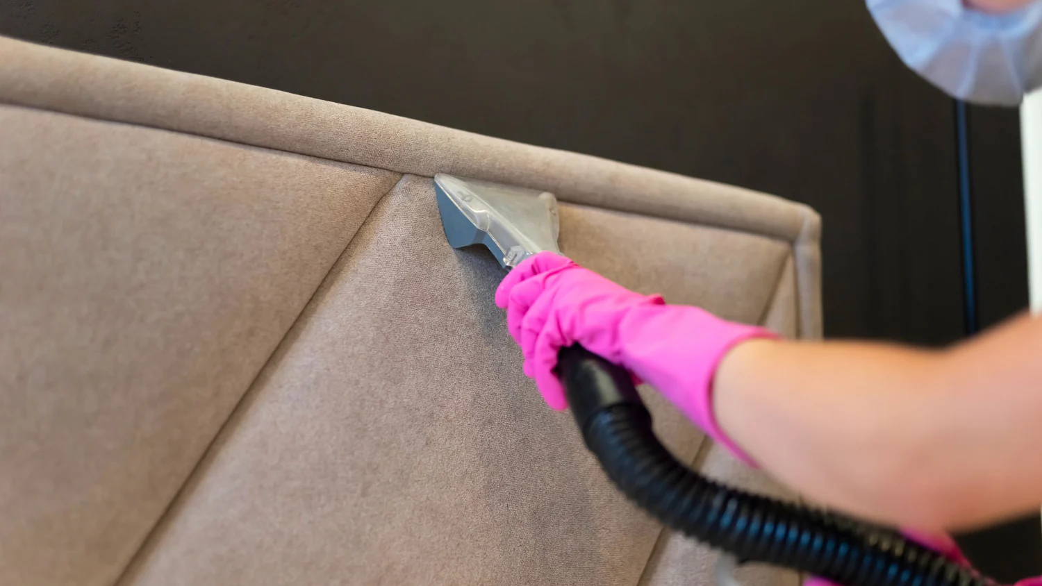 A tufted fabric headboard in a bright bedroom being gently vacuumed with an upholstery brush attachment, real home photo