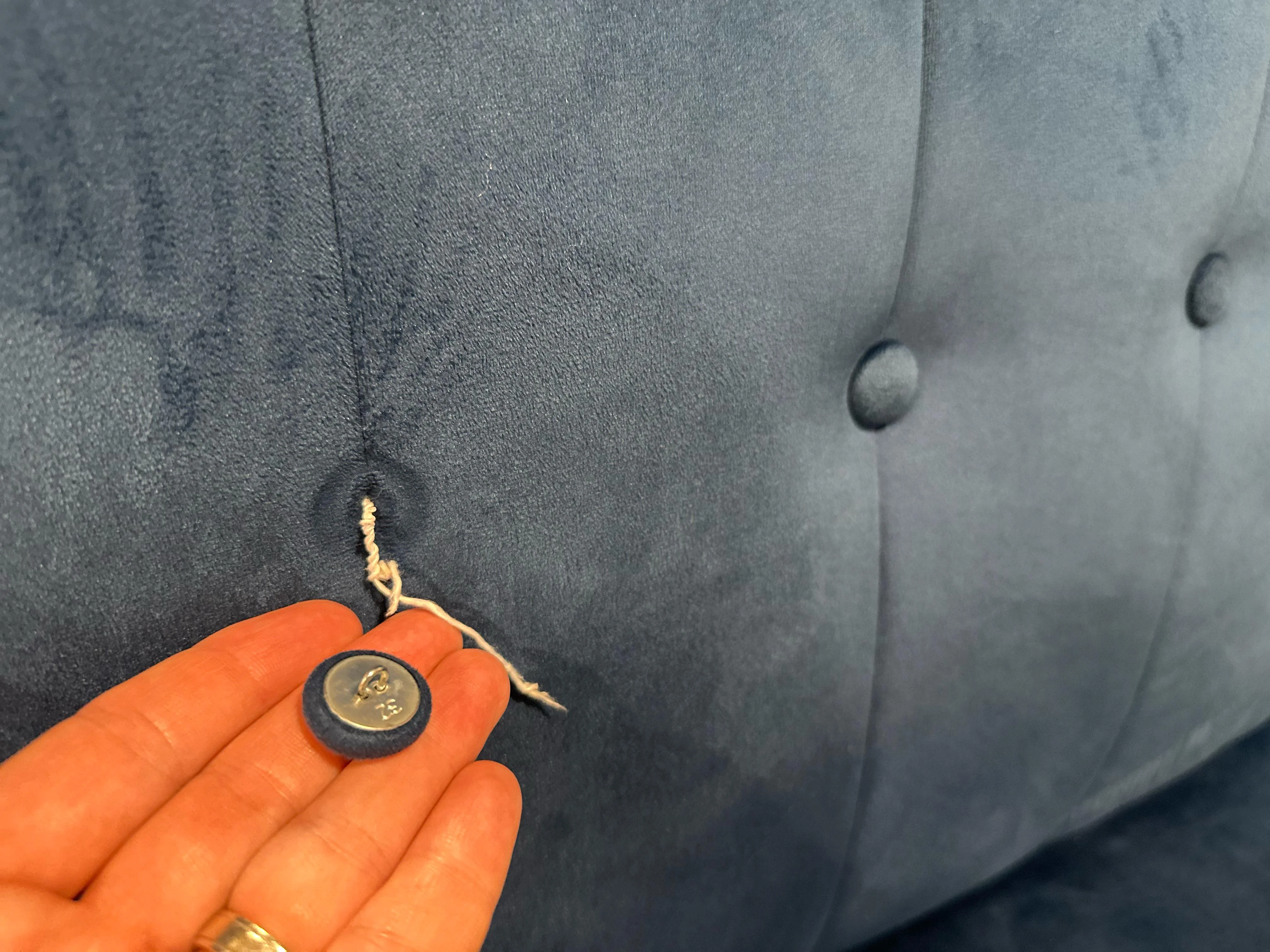 A tufting needle, braided upholstery twine, and a wide fender washer arranged on a wooden work surface next to a fabric-covered button