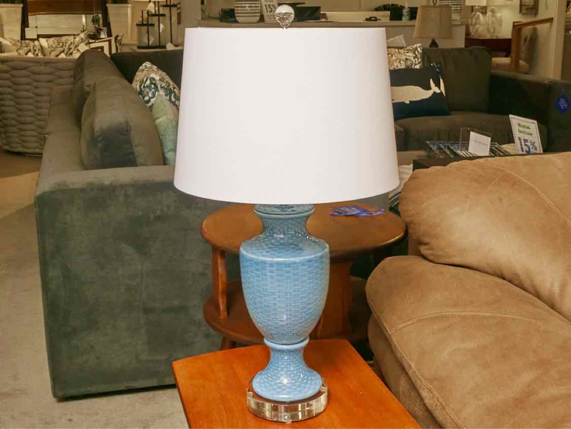 A vintage ceramic table lamp on a wooden side table with a softly textured white linen drum shade glowing warmly in a cozy living room, real photo
