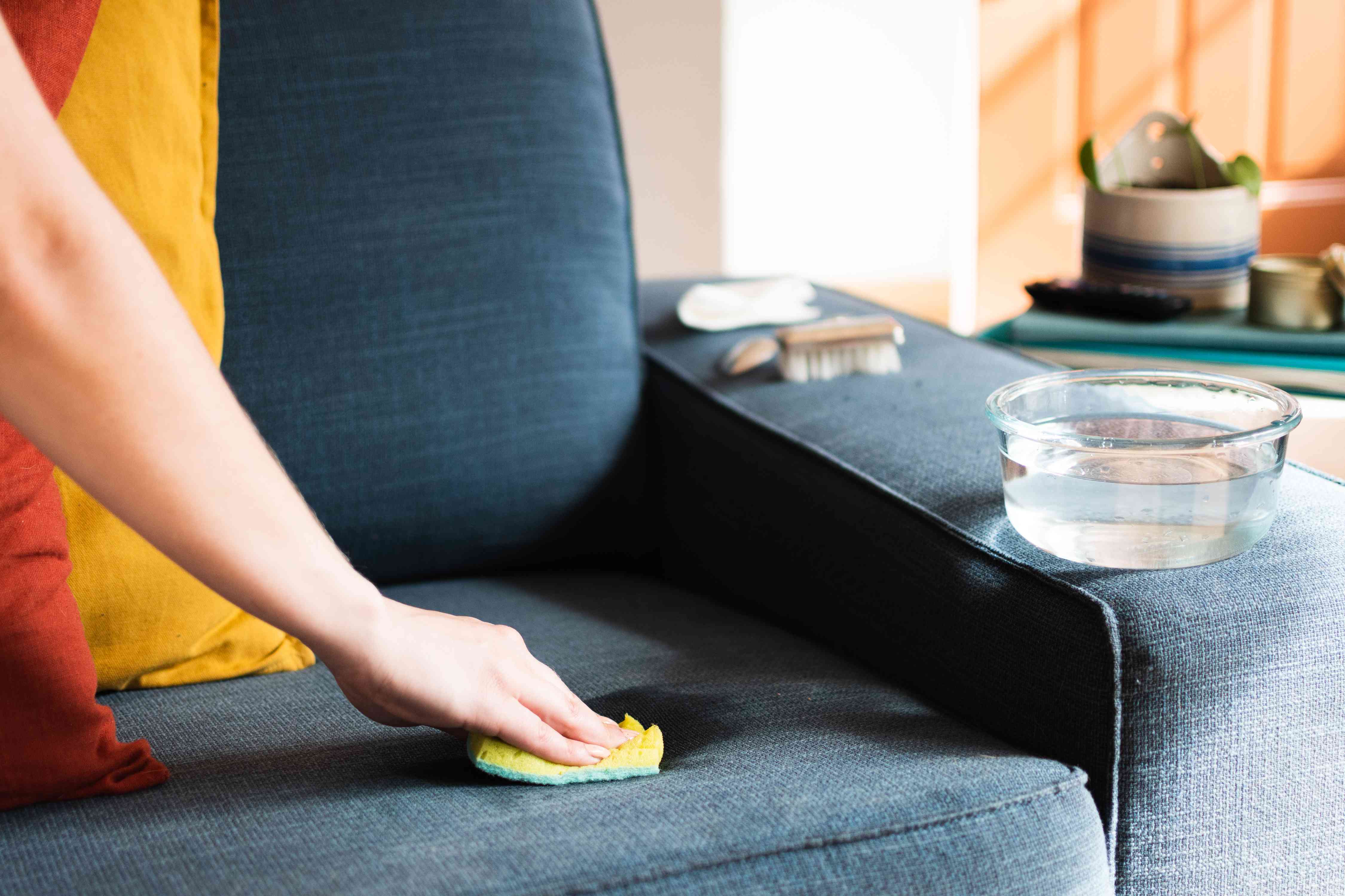 Close-up of hands gently blotting the arm of a neutral fabric accent chair with a white cloth and a small bowl of diluted dish soap nearby, realistic home photo