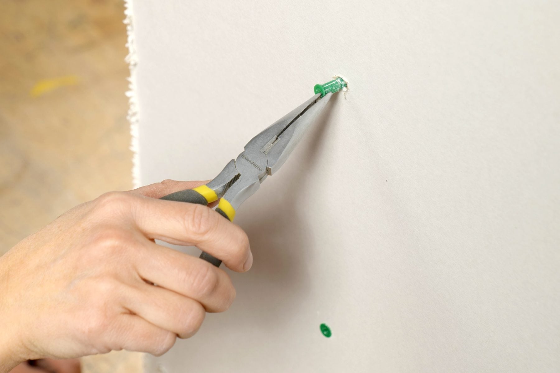 Close-up of hands using needle-nose pliers to gently pull a plastic drywall anchor from a painted wall, realistic home repair photo