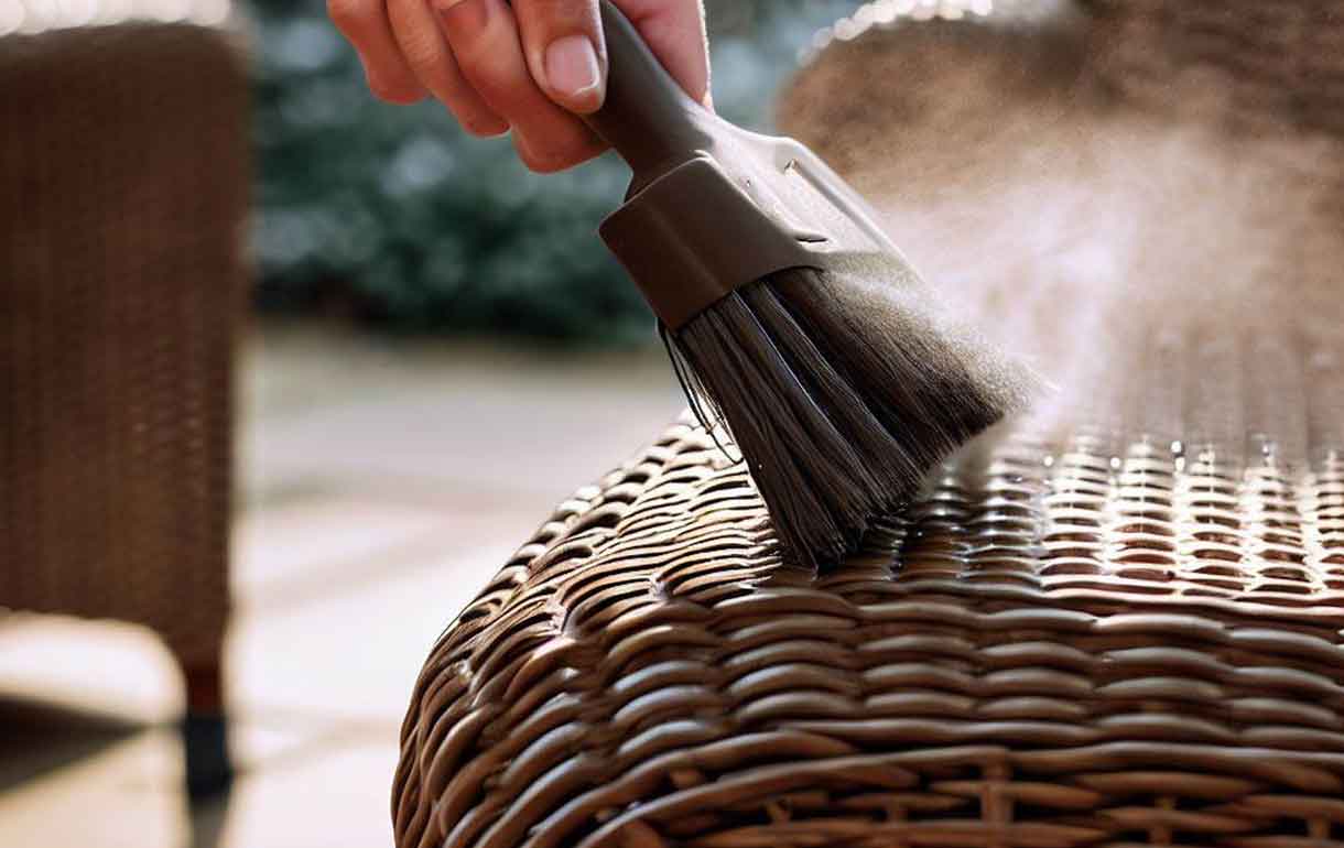 Close-up photo of a hand using a soft brush to dust debris out of the weave of an outdoor wicker chair, natural daylight