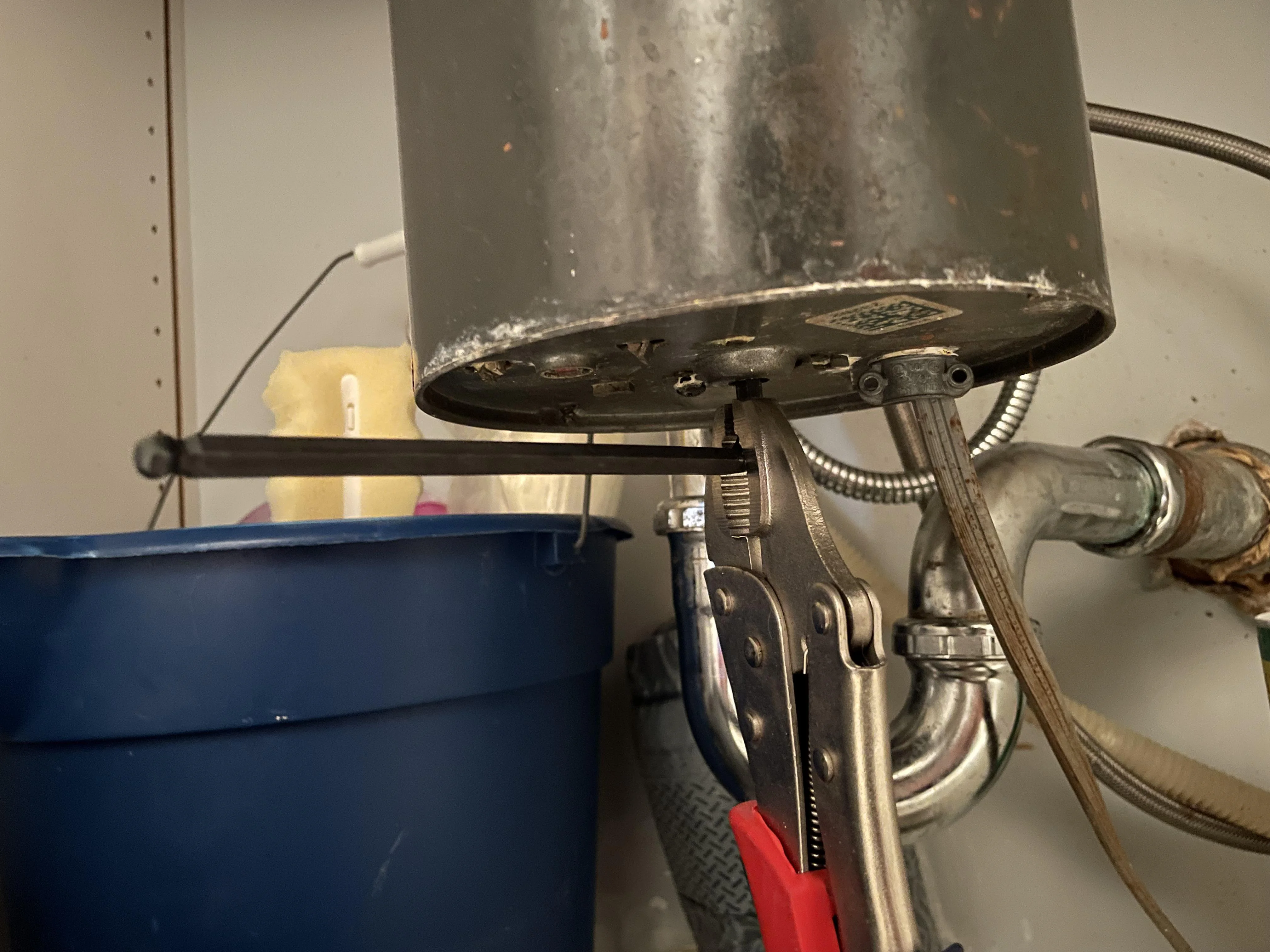 Close-up photo under a kitchen sink showing the bottom of a garbage disposal with an Allen wrench inserted into the hex socket, warm indoor lighting