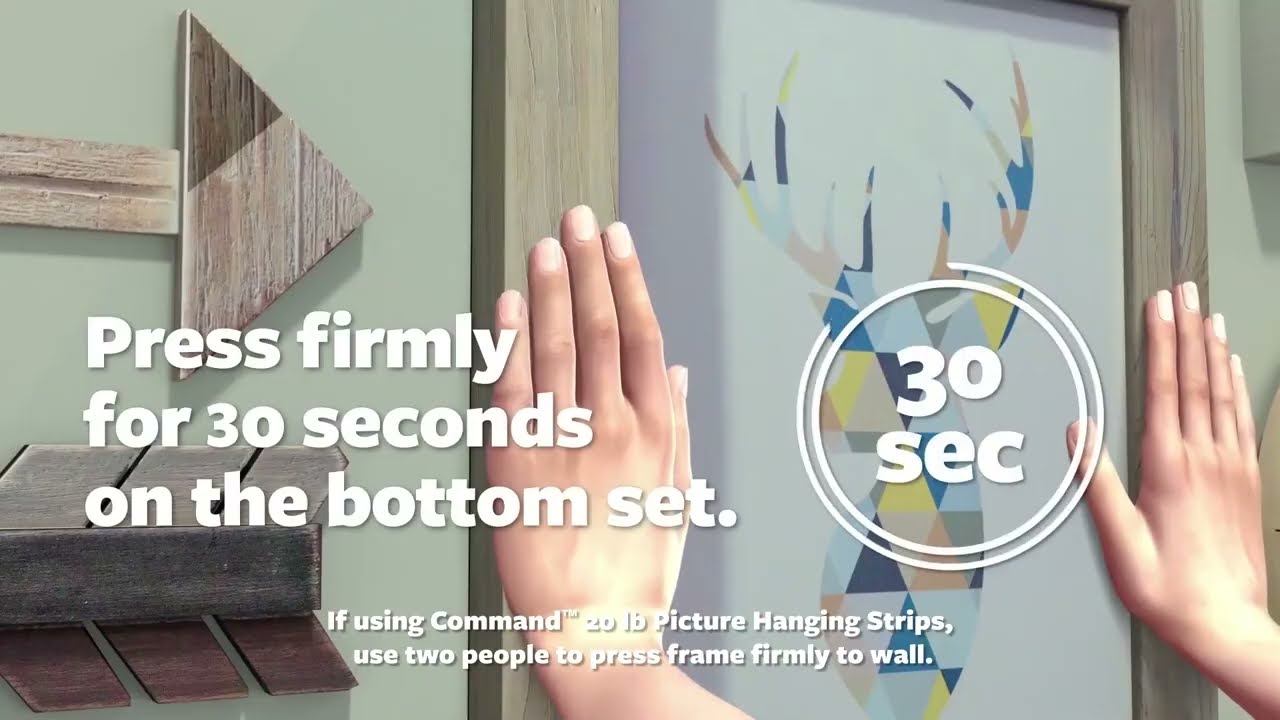 Hands pressing a framed print onto a wall using adhesive picture hanging strips in a bright apartment, real photograph