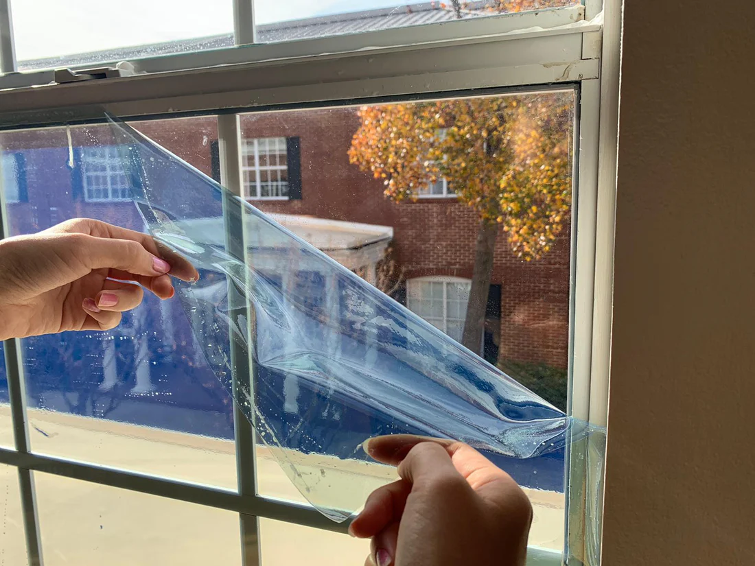 Hands using a small squeegee to smooth static cling privacy film on a clean apartment window, close-up real photo