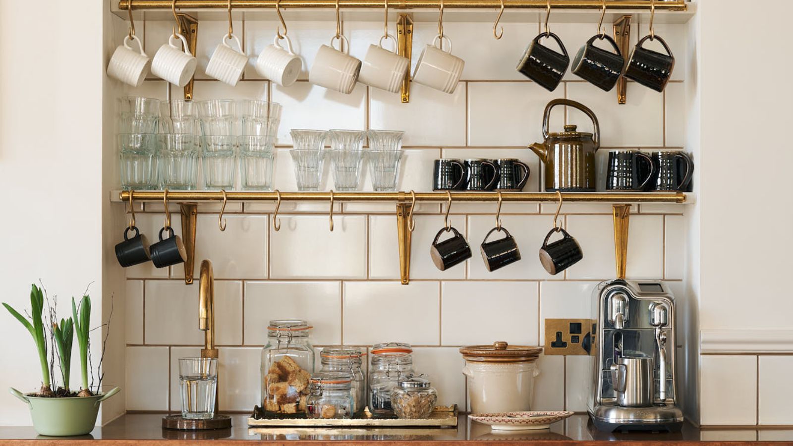 Kitchen coffee station on open shelves with matching mugs on hooks, a jar of coffee beans, a small tray with sugar and spoons, and a framed print leaning against the backsplash